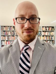 headshot of Christopher Brick, a bald man with red facial hair and black glasses, wearing a gray pinstripe suit with a pink undershirt and pink and blue striped tie, standing in front of bookshelves